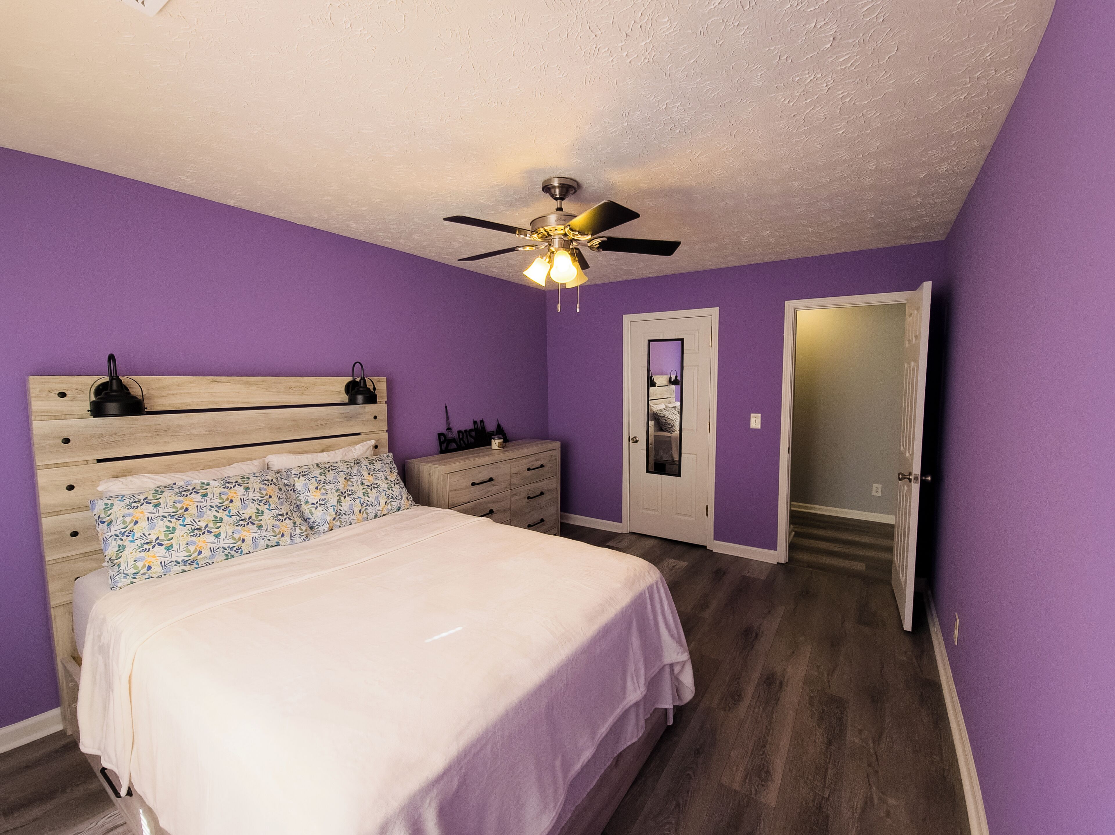 A bedroom with a bed, a nightstand, a dresser, a ceiling fan, and a door.