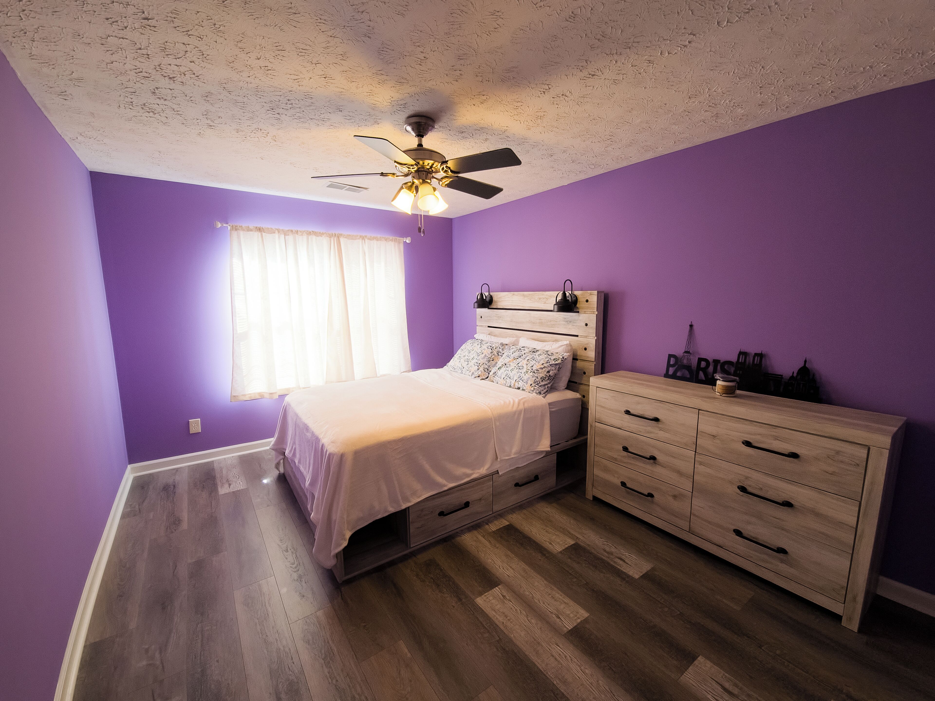 A bedroom with a bed, a dresser, a ceiling fan, and a window with curtains.