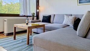 Een gezellige woonkamer met een bank, een houten tafel met een fles en glazen, een radiator en een raam met gordijnen.