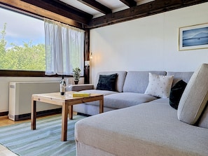 A cozy living room with a sofa, a wooden table with a bottle and glasses, a radiator, and a window with curtains. - Ferienpark Vorauf by Interhome (Siegsdorf)