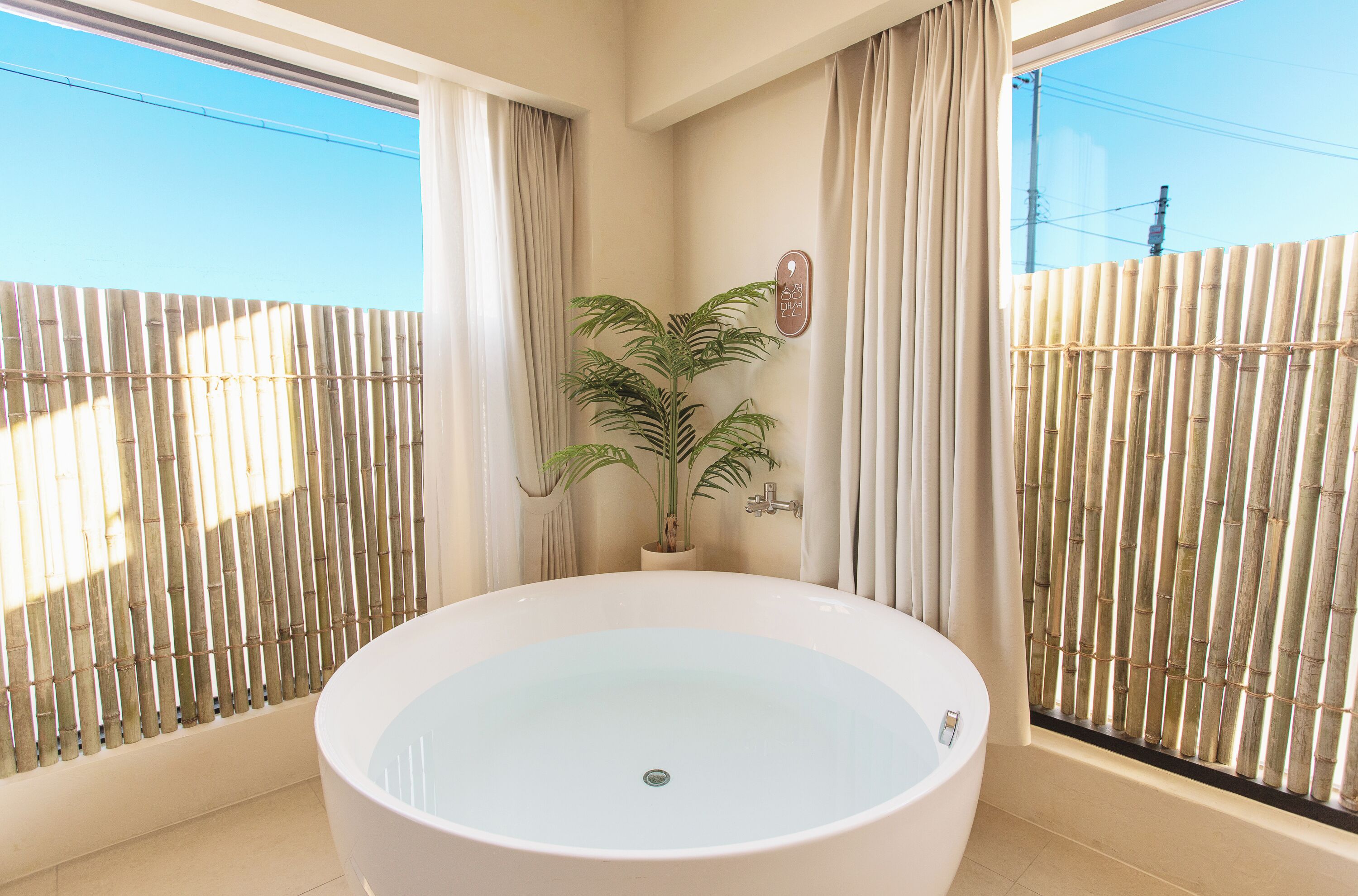 A bathroom with a large bathtub, a potted plant, and a bamboo fence outside the window.