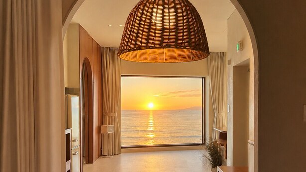 A hotel room with a large window, a bed, a hanging lamp, and a view of the sunset over the water.
