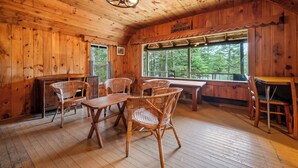 A wooden cabin interior with a dining area, wicker chairs, a wooden table, and a large window offering a view of trees.