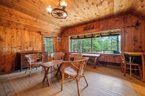 A wooden cabin interior with a dining area, wicker chairs, a wooden table, and a large window offering a view of trees.