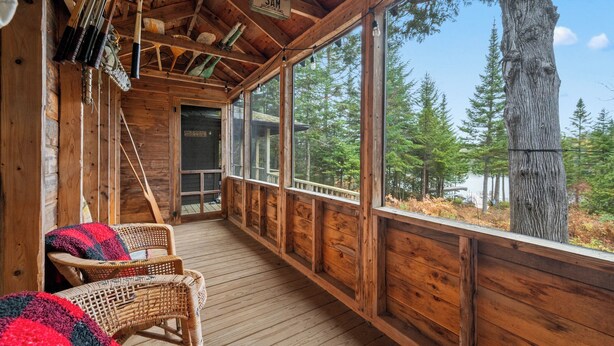 A wooden porch with wicker chairs, a sign reading 'SAM,' and a view of trees and a lake.