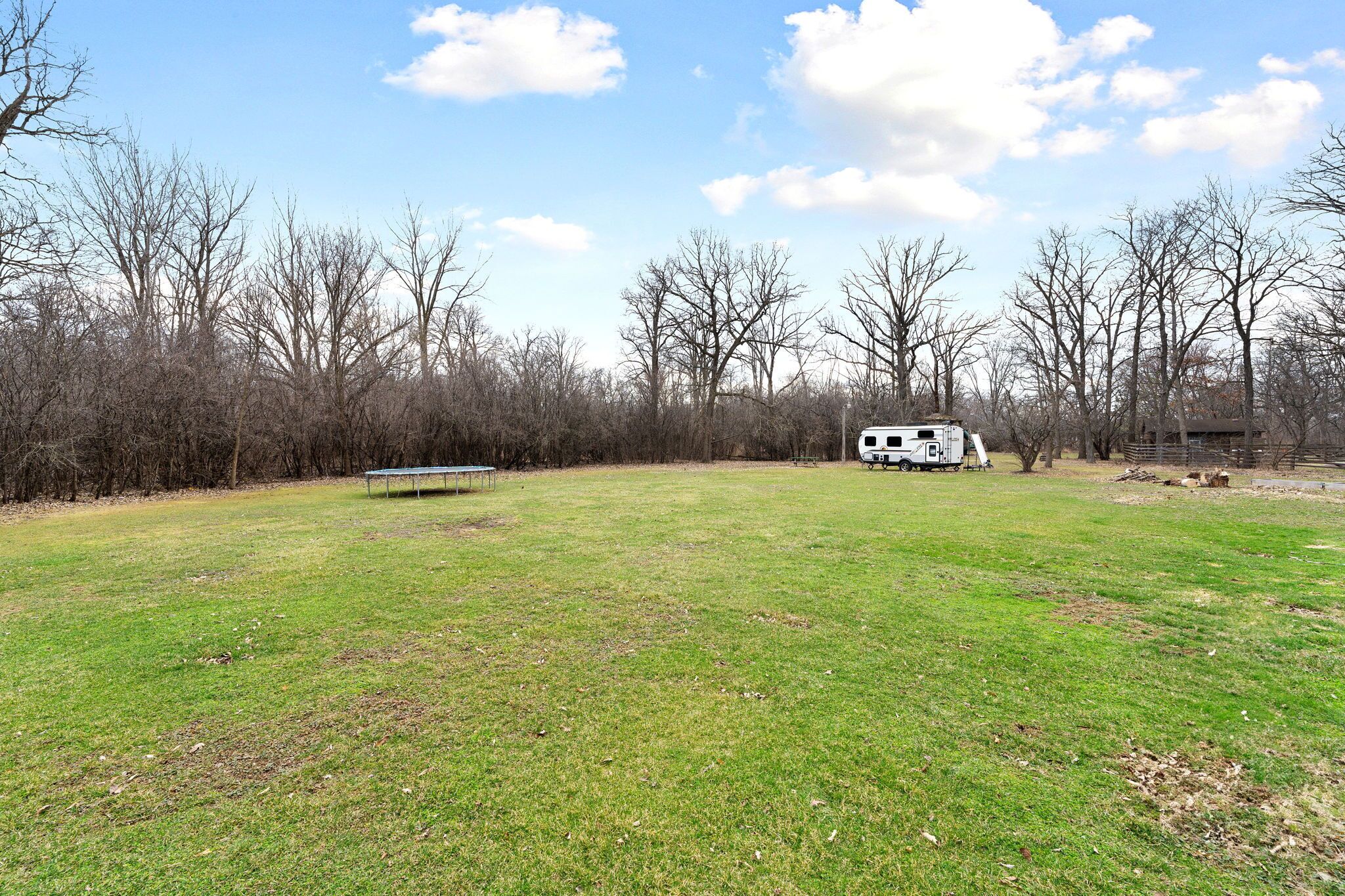 A grassy field with a bench, a white RV, and leafless trees.