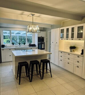 A modern kitchen with a central island, bar stools, white cabinetry, and a view through large windows. - Boutique farm-stay in The Southern Highlands with pool. Family + pet friendly. (Woodlands)