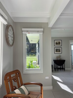Ein Zimmer mit Holzstuhl, einem Fenster mit Gartenblick, einer Wanduhr und einem gerahmten Bild.