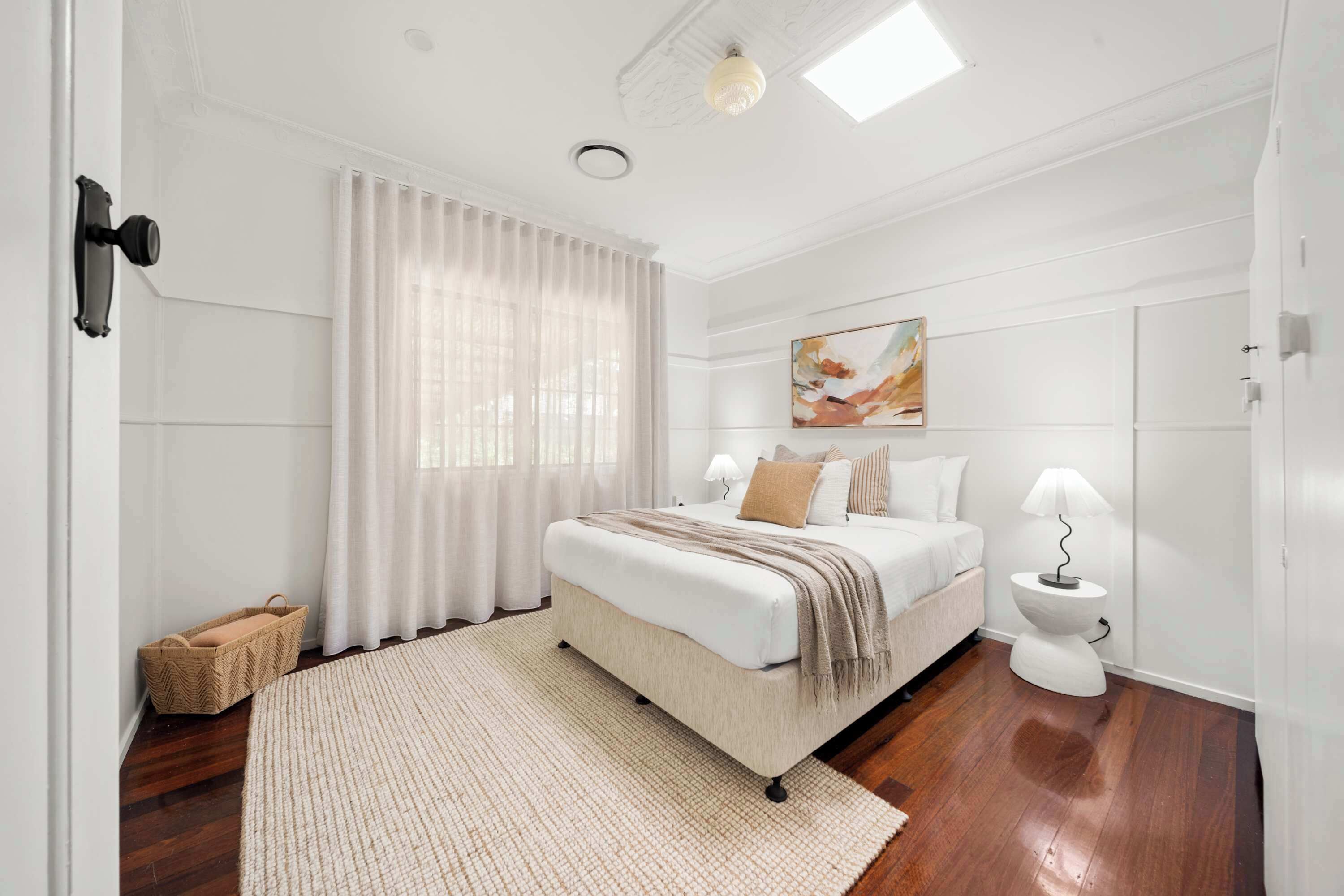 A bedroom with a bed, bedside lamps, a window with curtains, a rug, and a painting on the wall.
