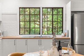 Eine Küche mit weißen Schränken, einem Edelstahlkühlschrank, einem Fenster mit Blick auf Grünflächen und einem Esstisch, gedeckt mit Gläsern und einer Flasche.