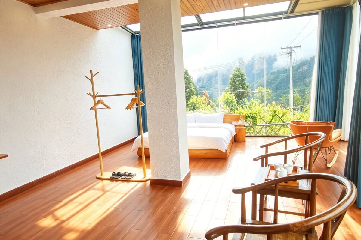 A spacious room with wooden flooring, a bed, a seating area with chairs, a coat rack, and a large window with a view of greenery and hills.