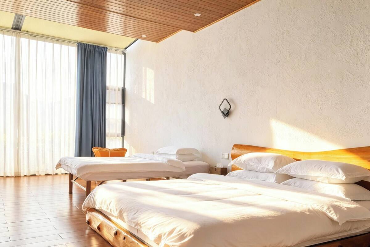 A hotel room with two beds, a wooden ceiling, a large window with curtains, and a wall-mounted light fixture.