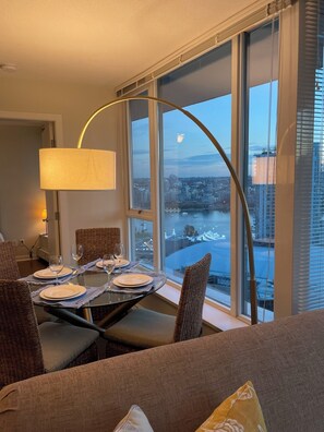 Ein Essbereich mit Tisch für vier Personen, großen Fenstern mit Blick auf eine Stadtlandschaft und einer modernen Lampe.