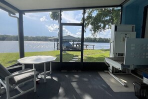 A room with a large window overlooking a lake, a table, a chair, and a grill area.
