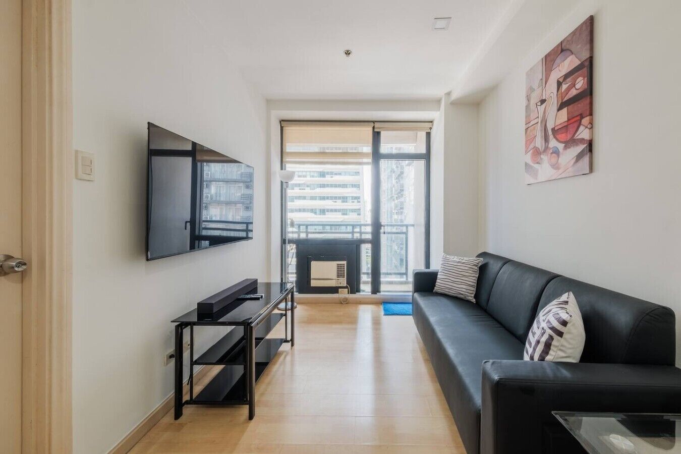 Een moderne woonkamer met een zwarte bank, een aan de muur bevestigde televisie, een salontafel en een groot raam met uitzicht op gebouwen.