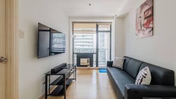 Een moderne woonkamer met een zwarte bank, een aan de muur bevestigde televisie, een salontafel en een groot raam met uitzicht op gebouwen.