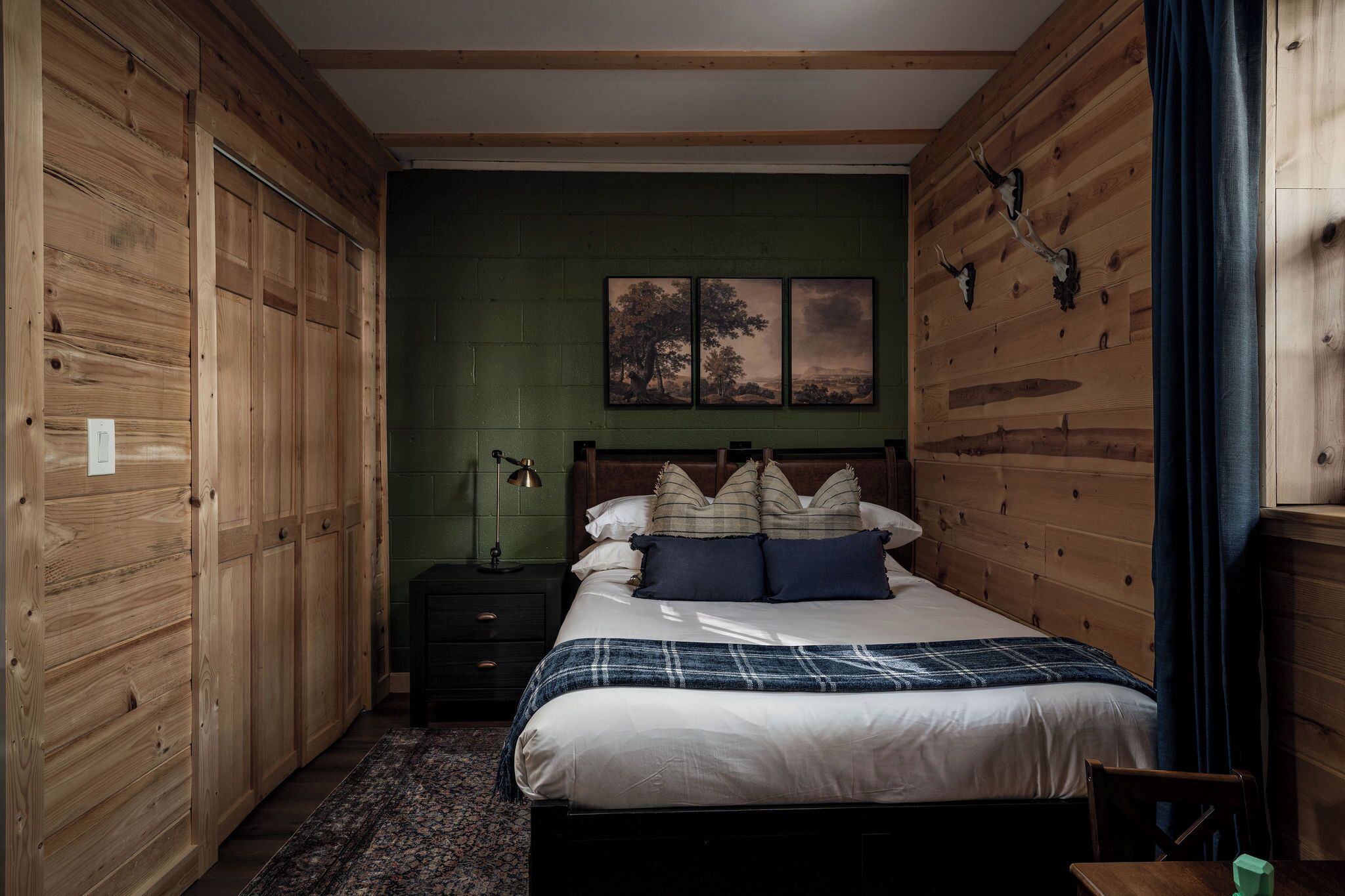 A bedroom with a wooden headboard, a bed with white and blue bedding, a nightstand with a lamp, and a green wall with artwork.