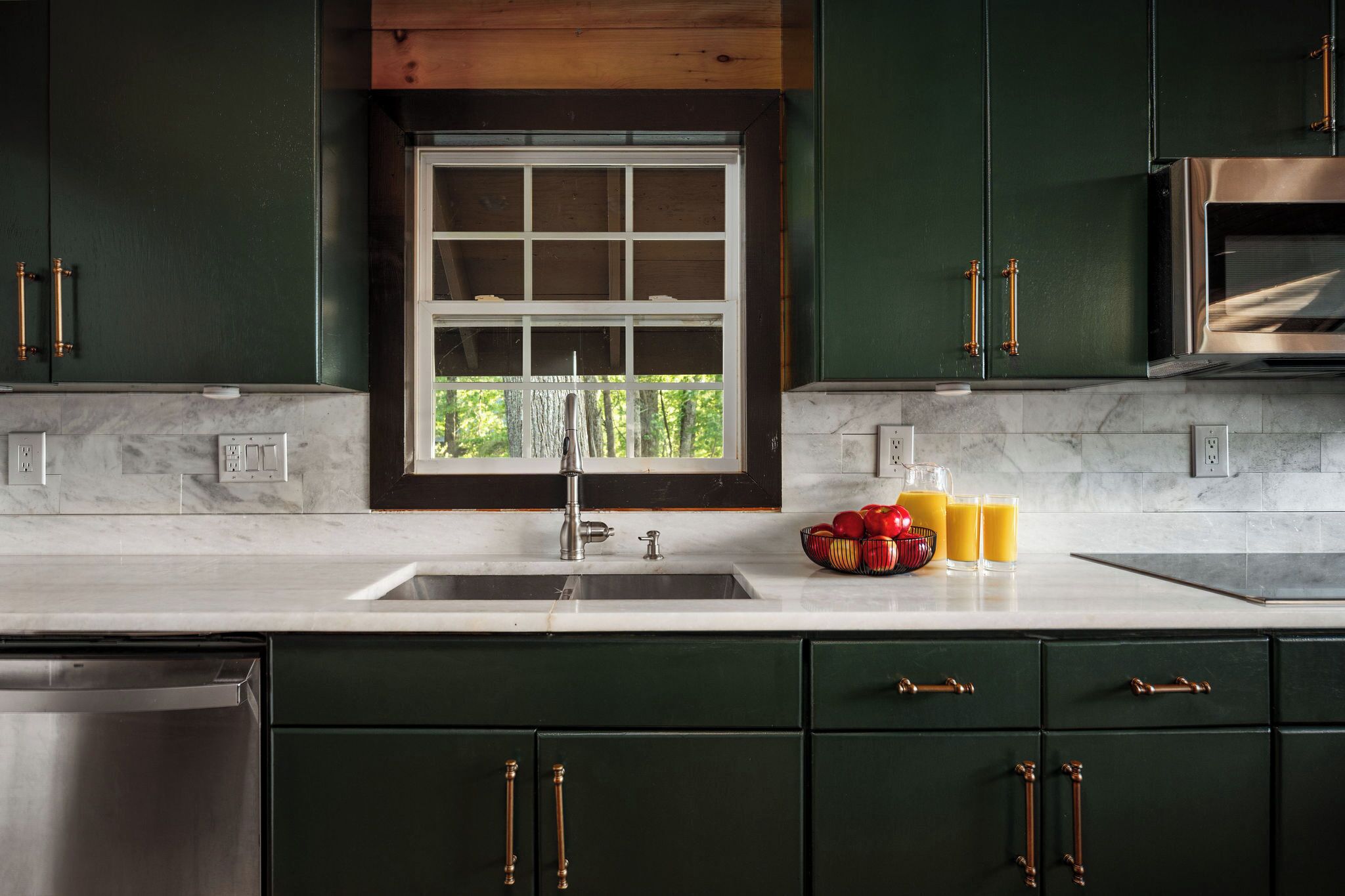 A modern kitchen with dark green cabinets, a stainless steel dishwasher, a double sink, and a window with a view of greenery.