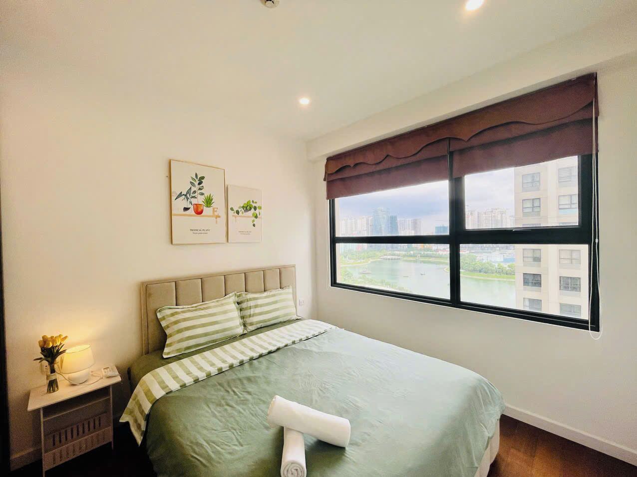 A neatly arranged bedroom with a bed, a bedside table with a lamp, and a window with a view of buildings and a body of water.