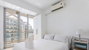 A modern hotel room with a bed, a nightstand, a lamp, and a view of buildings through large windows. - StayHere 2BR Beach Tropical City Getaway Azure (Parañaque)