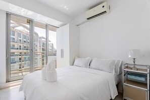 A modern hotel room with a bed, a nightstand, a lamp, and a view of buildings through large windows. - StayHere 2BR Beach Tropical City Getaway Azure (Parañaque)