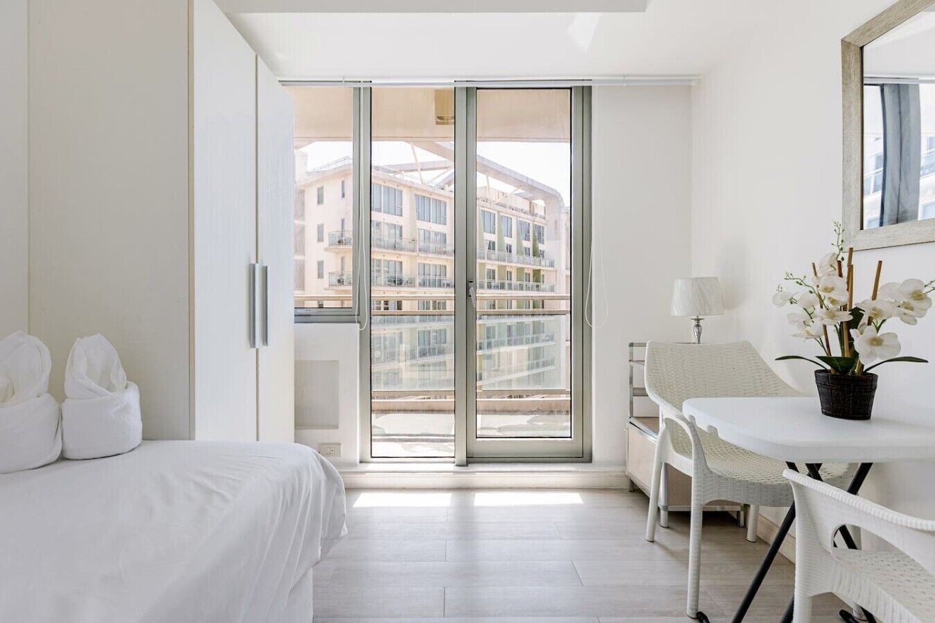 Hotellihuone, jossa on sänky, työpöytä tuolilla, ruukkukasvi ja liukuovet, jotka johtavat parvekkeelle, jolta on näkymä rakennuksiin.