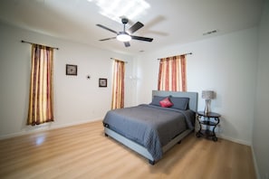 Un dormitorio con cama, ventilador de techo, cortinas, una mesita de noche y una lámpara.