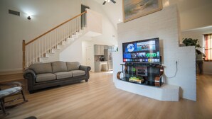 A living room with a sofa, a TV mounted on a brick wall, a staircase, and a kitchen in the background. - A Quiet Lakefront Retreat for Relaxed Getaways (Oak Point)