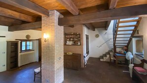 Um interior rústico com vigas de madeira aparentes, lareira de tijolos, escada e prateleira com utensílios de cozinha.