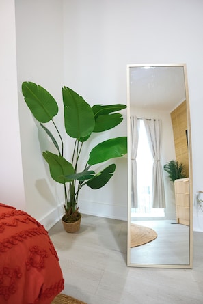 Ein Zimmer mit einem großen Spiegel, einer Topfpflanze, einem roten Kissen und einem Holzkasten.