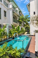 A swimming pool with a glass barrier, surrounded by lush greenery and a white building with balconies.