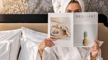 A person in a white bathrobe sitting on a bed with a book titled 'BRILLIANT JOURNAL' and a tray of pastries.