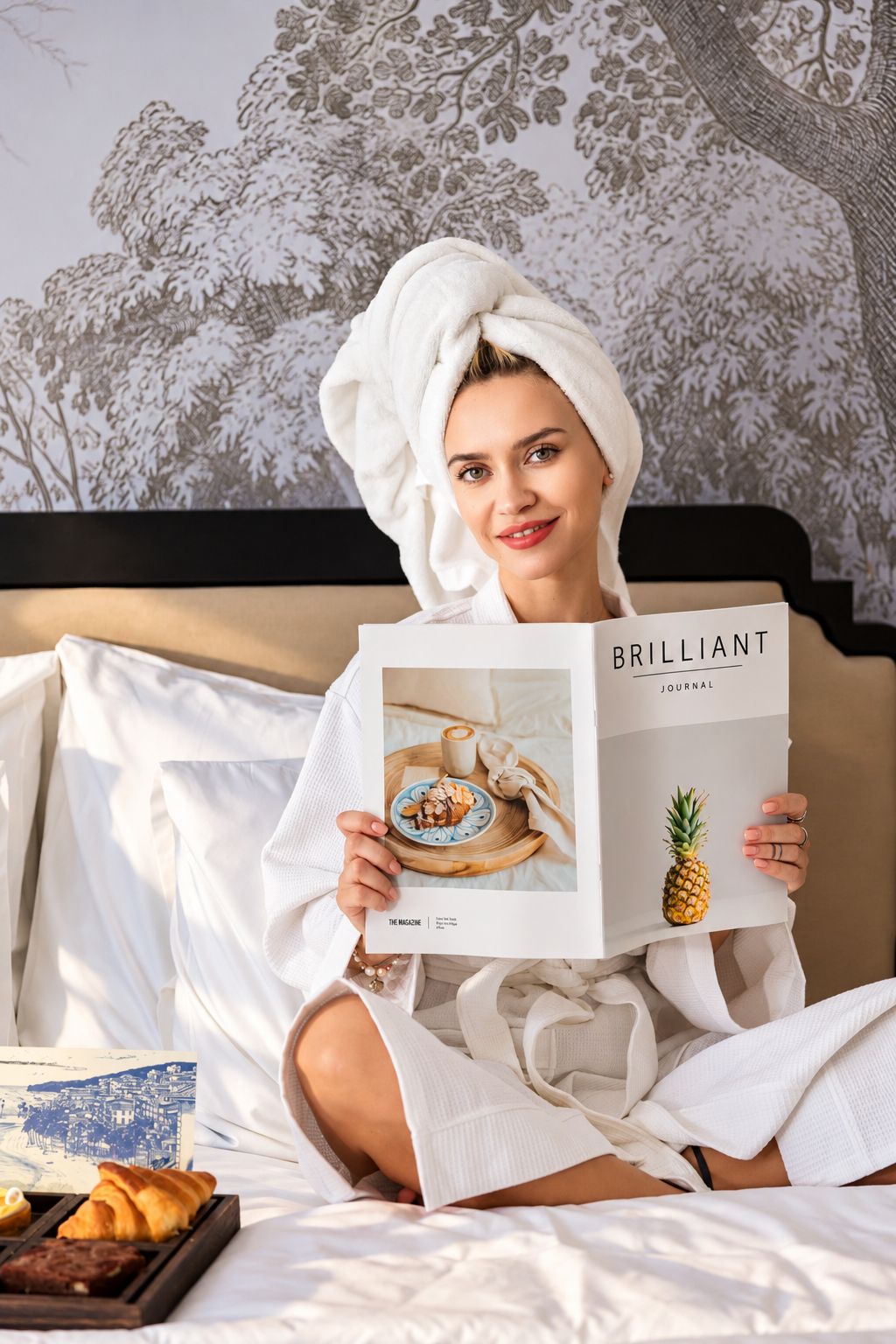 A person sitting on a bed with a book titled 'BRILLIANT JOURNAL' and a tray with pastries.