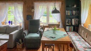Una sala de estar acogedora con un sofá, una butaca, una mesa de comedor con un banco, una estantería y un ventanal con cortinas.