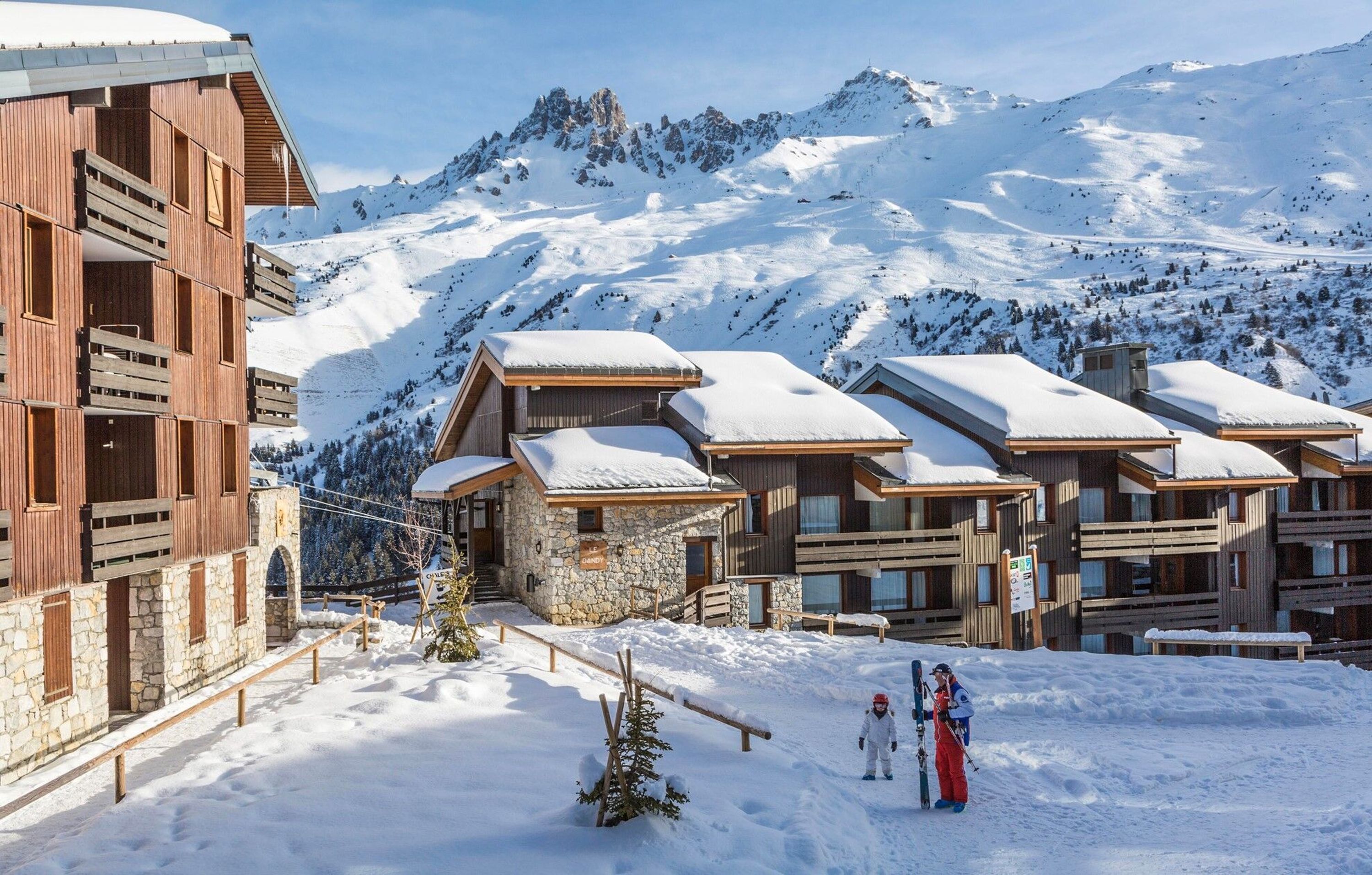 A snow-covered lodge with stone and wooden buildings, ski slopes in the background, and a clear sky.