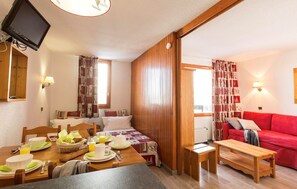 Ein gemütliches Zimmer mit Frühstückstisch, einem Bett mit Bettdecke, einem roten Sofa und einem an der Wand befestigten Fernseher.
