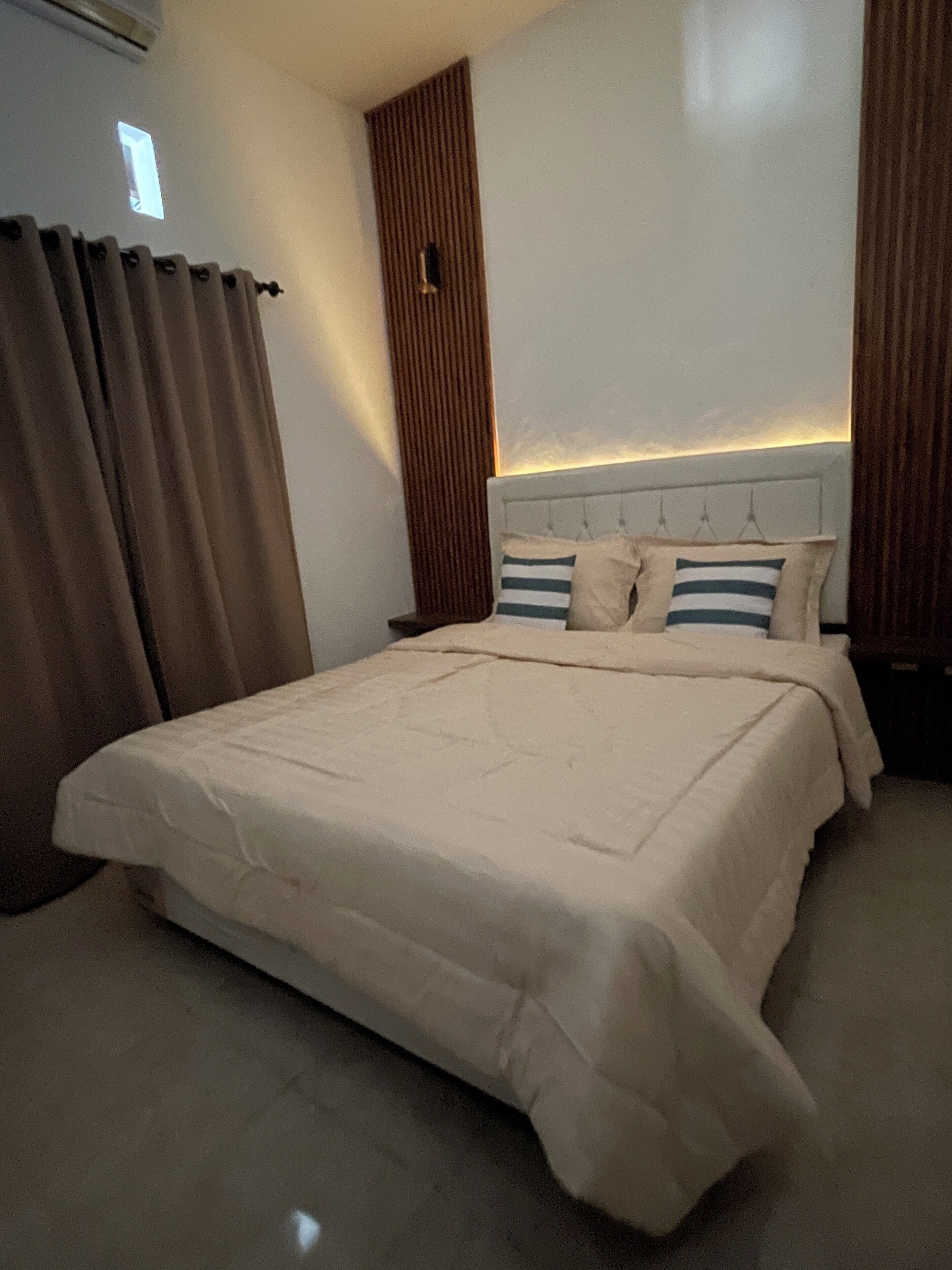 Une chambre à coucher avec un grand lit, une tête de lit dotée d’un éclairage intégré, une table de chevet et une fenêtre avec des rideaux.