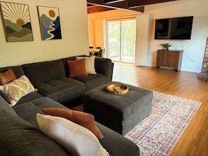 Un salon moderne avec un canapé d’angle, une table d’appoint en bois et un grand téléviseur à écran plat fixé au mur.