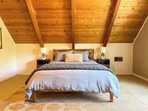 A bedroom with a wooden ceiling, a bed with a grey duvet, two bedside tables with lamps, and a patterned rug.