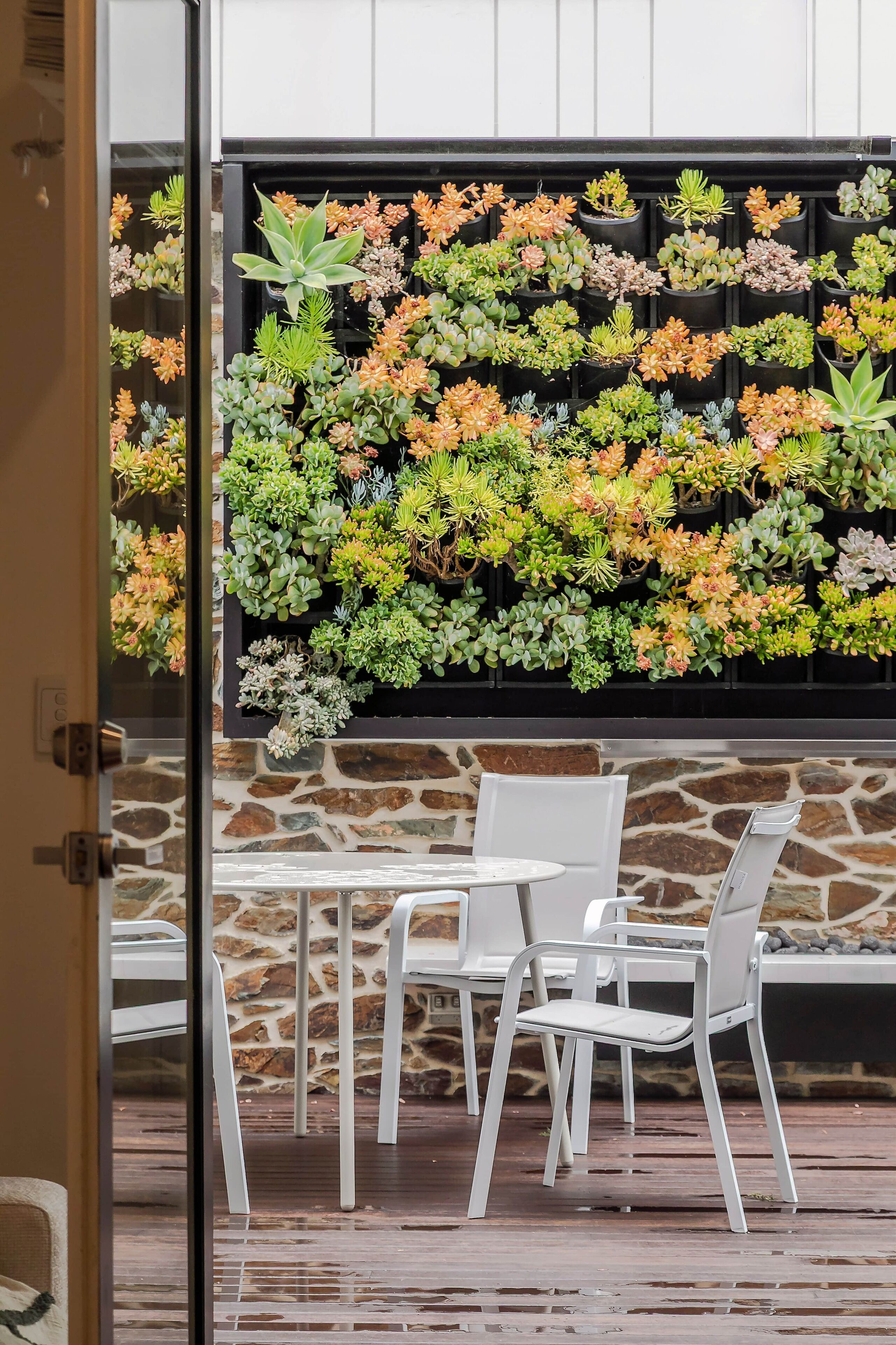 A patio area with white chairs and a table, a stone wall, and a wall of various succulents.