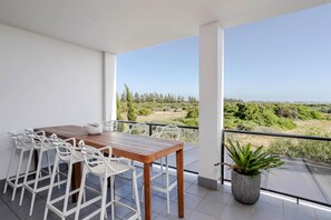 A balcony with a wooden table and white chairs, a potted plant, and a view of a green landscape.