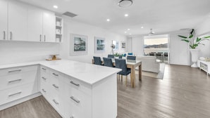 A modern kitchen with white cabinetry, a dining area with a wooden table and chairs, and a view of the outdoors.