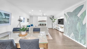 A modern living room with a glass dining table, grey upholstered chairs, a white sofa, and a large TV.