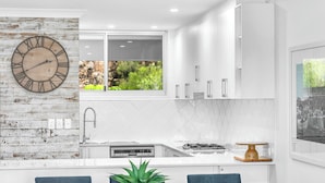 A modern kitchen with white cabinetry, a wooden clock, a window with a view of greenery, and a sink.