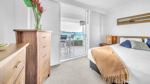 A hotel room with a bed, a wooden dresser, a nightstand, a lamp, and a view of the balcony.