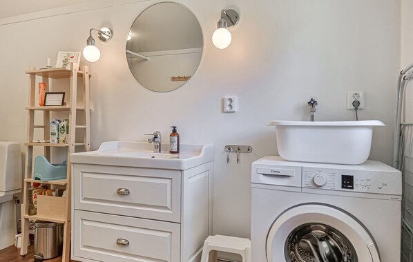 A compact bathroom with a sink, mirror, wall-mounted lights, a washing machine, and a small shelf with toiletries. - Beach front home in Bullaren with house sea view (Bullaren)