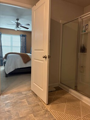 Une chambre à coucher comprenant un lit, un ventilateur de plafond et une porte donnant sur une salle de bain avec une douche à parois de verre.