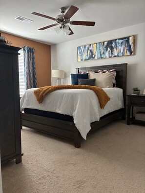 Un dormitorio con cama, ventilador de techo, un cuadro, lámpara y ventana con cortinas.