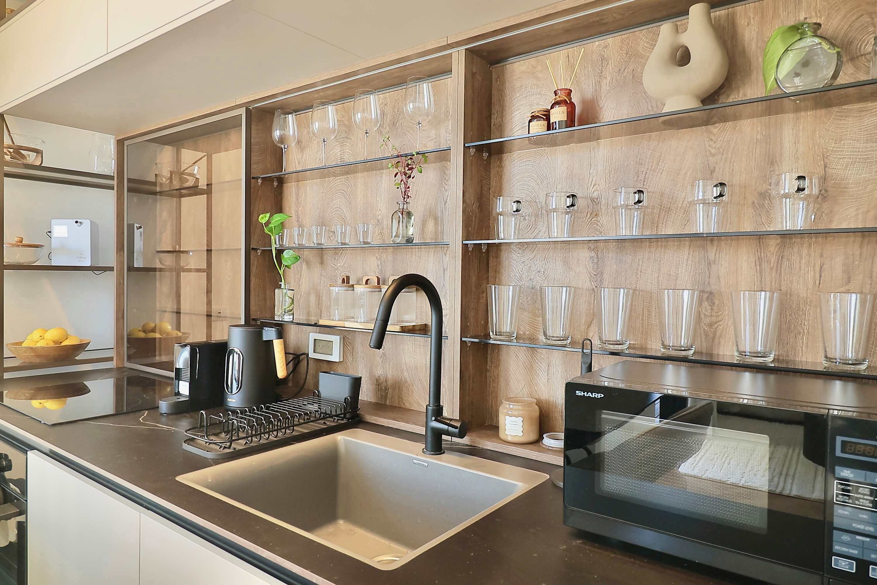 Une cuisine moderne dotée d’un évier en acier inoxydable, d’un micro-ondes noir et d’un mur en bois avec des étagères où sont rangés de la verrerie et des bocaux.