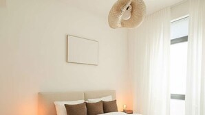 Une chambre à coucher comprenant un lit, des tables de chevet, un luminaire suspendu, une fenêtre avec des rideaux et un tableau encadré accroché au mur.